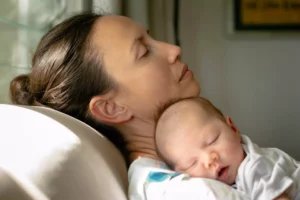 A woman leans her head back on a couch, while cuddling a newborn baby on her shoulder.