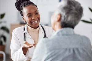 A close up of the back of a patient's head, facing a doctor wearing a white lab coat with a stethoscope around her neck. They could be discussing healthcare pricing.