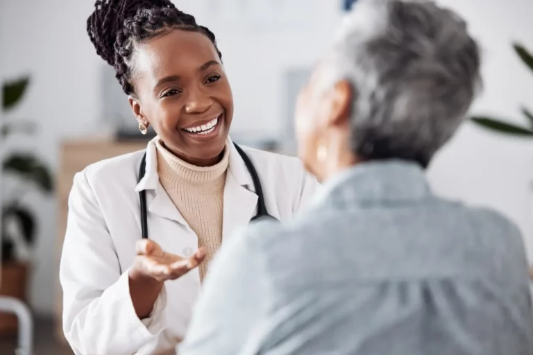 A close up of the back of a patient's head, facing a doctor wearing a white lab coat with a stethoscope around her neck. They could be discussing healthcare pricing.